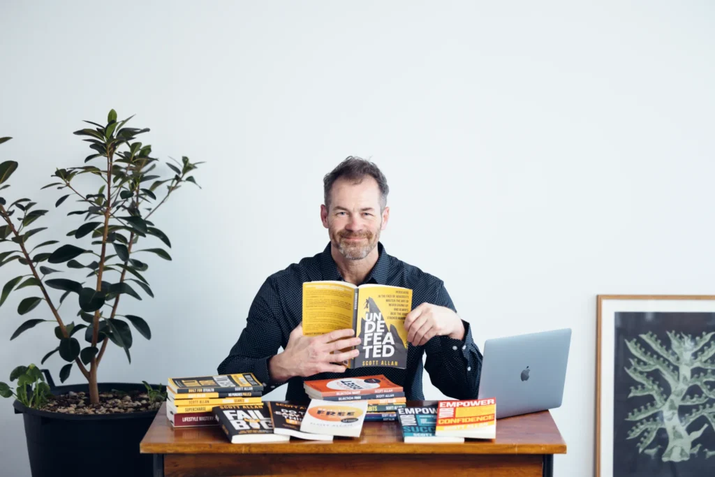 scott allan about the author The author Scott Allan sitting at a desk, holding one of his books and with more of his books scattered on the desk.